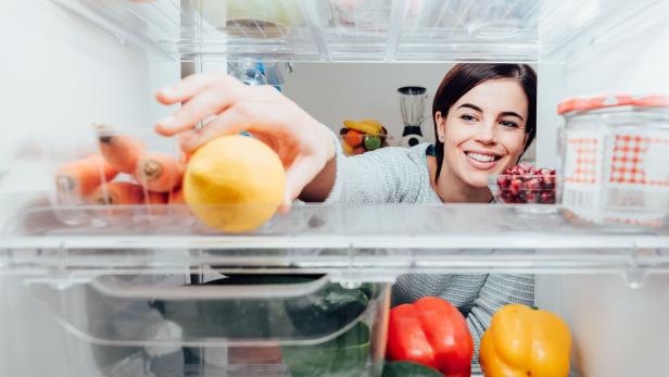 Eine Frau greift lächelnd nach einer Zitrone in einem Kühlschrank, der mit Gemüse und Obst gefüllt ist.